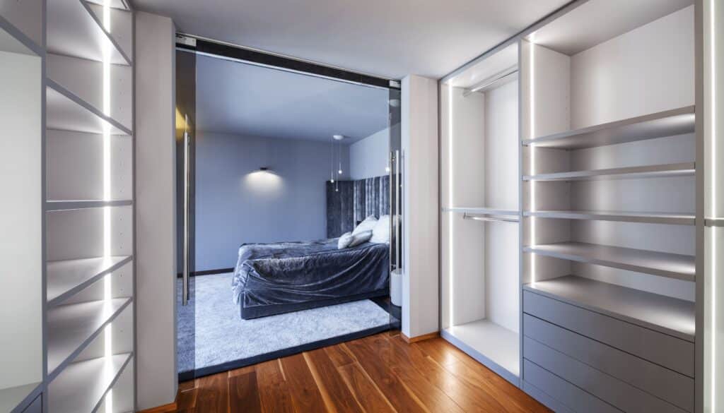 Moderne dressing avec étagères éclairées, tiroirs gris et tringle, séparé d'une chambre élégante par une porte vitrée. Lit gris, sol parquet/moquette.