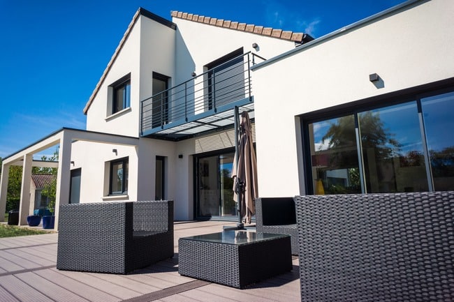 Maison moderne blanche avec terrasse en bois, mobilier de jardin gris, et balcon. Ciel bleu éclatant.