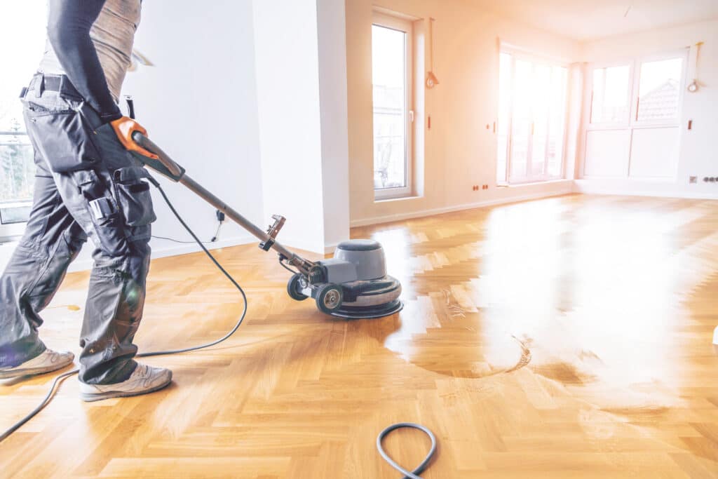 Ouvrier professionnel vitrifiant un parquet en bois clair à chevrons avec une polisseuse. La zone traitée brille sous la lumière des fenêtres.