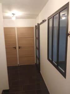 Couloir avec sol en carrelage foncé, deux portes en bois clair modernes, une verrière style atelier coulissante et un plafonnier simple.