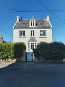 Maison deux étages couleur crème sous ciel bleu clair, avec toit en ardoise, lucarne, cheminée et échelle de toit. Panneau 'DEVIS GO' à l'entrée.
