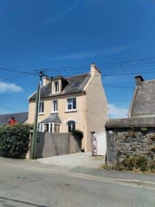 Maison beige à deux étages avec lucarnes sous un ciel bleu clair, poteau électrique et échelle sur le toit. Numéro de maison '14'.