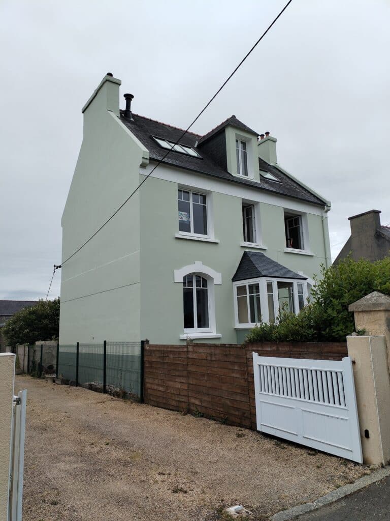 Maison ancienne repeinte en vert pâle avec toiture en ardoise, lucarne, et petit bow-window. Portail blanc à droite.