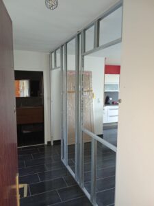 Structure métallique de cloison sèche (montants Stil) en cours d'installation, vue depuis l'entrée avec carrelage sombre et accès à une salle de bain et une cuisine.
