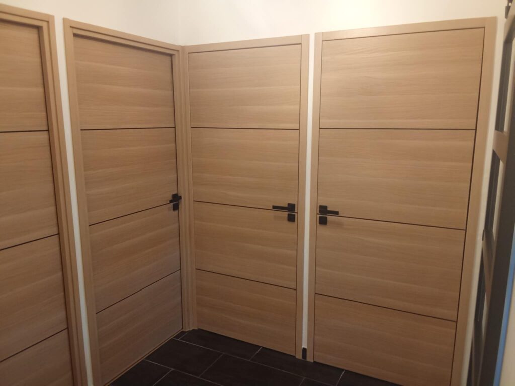 Quatre portes modernes en placage bois clair avec rainures noires horizontales et poignées noires plates, sur sol carrelé sombre.